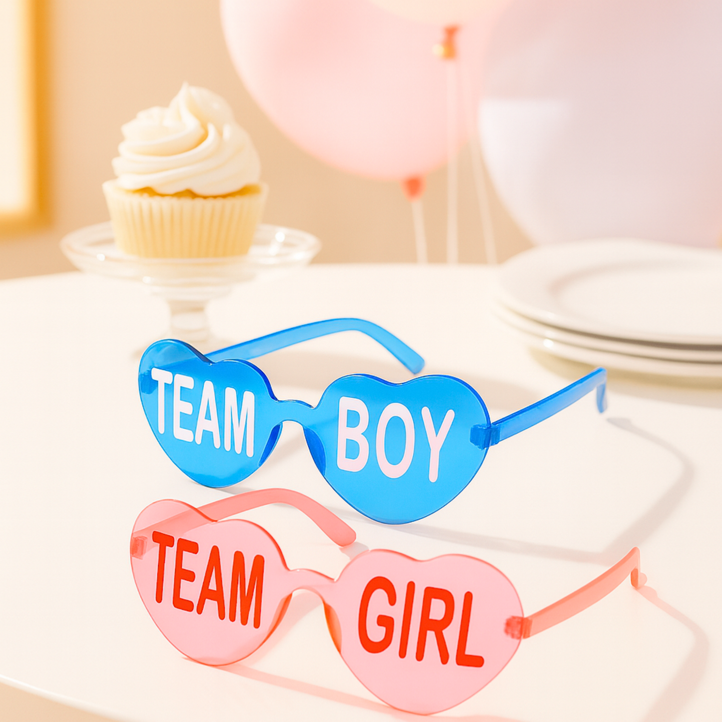 Blue Team Boy and pink Team Girl heart-shaped glasses styled as gender reveal party photo props on a table with cupcake and pastel balloons.